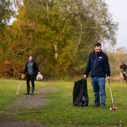 A man and a woman holding litter picks and bin bags in a countryside location with trees in the background