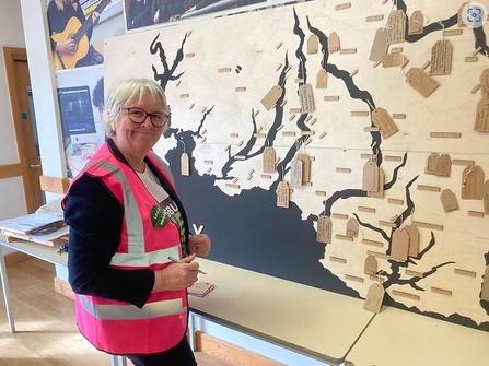 A woman with short grey hair wearing a bright pink hi-vis jacket standing in front f a map showing rivers in South Devon