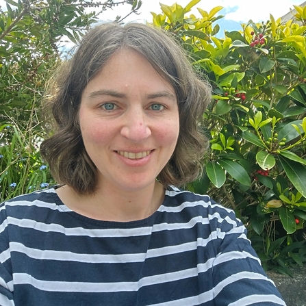 A woman with short brown hair and blue eyes, wearing a navy and white striped top, standing in front of a hedge