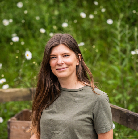 A woman with long brown hair, wearing a khaki green T-shirt, standing in front of a fence post in a field with white flowers in the background