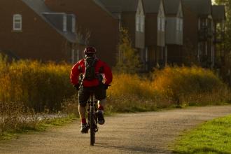 Man riding bike down path by a river with houses in background.