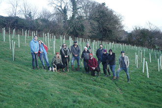 A group of people standing in a field with newly planting trees wrapped in protective plastic