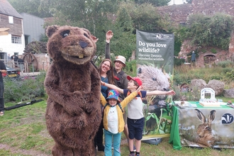 Family of two adults and two children stretching their arms next to large beaver costume in garden 