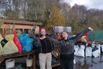 A man and a woman with hands in the air looking happy standing in front of tables filled with tree saplings at a tree hub