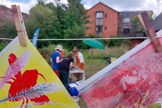 Colourful printed artwork pegged to a clothesline in the sunshine, with people and a sun umbrella in the background
