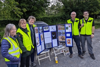 A group of batwalk guides, standing next to an information display, wearing high vis jackets with bat emblems
