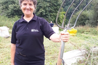 A person smiling stands holding a pitchfork with a bag of grass and branches beside them.