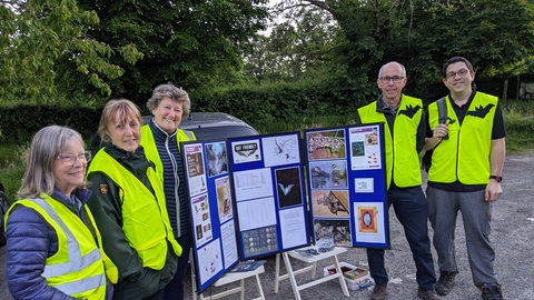 A group of batwalk guides, standing next to an information display, wearing high vis jackets with bat emblems