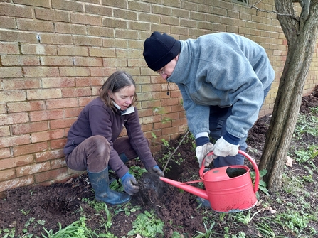 A woman crouched down in a plant bed and a person with a hat on holding a watering can