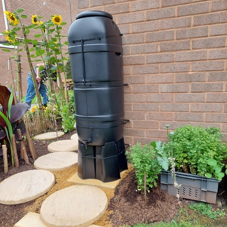 Water butt next to a building and sunflowers in the background