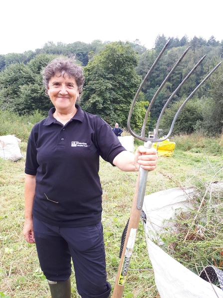 A person smiling stands holding a pitchfork with a bag of grass and branches beside them.
