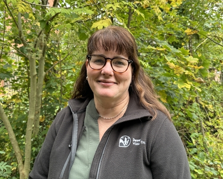 Angela Mather smiles at the camera with autumnal trees behind her