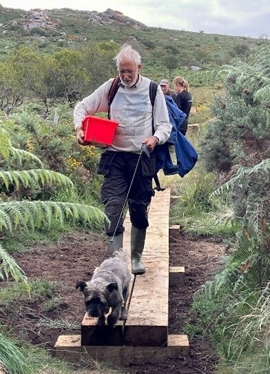 John walking along a boardwalk, gear in hand, wellies on and a dog leading the way