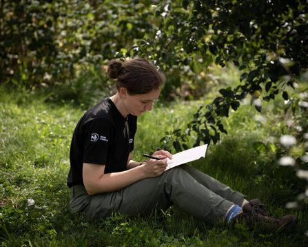 Jaz conducting a survey whilst sitting on the grass under a tree writing notes on paper