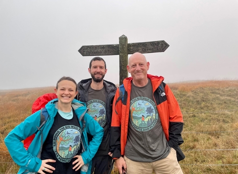 A woman and two men stood in front of a sign post on the Pennine way. The weather is misty.