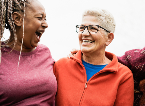 Two women, one black with braids and one white with short grey hair and glasses, laughing and smiling together